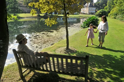 Visiteurs dans le jardin, homme assis sur un banc au bord de l'eau, un femme et sa fille avance vers lui.