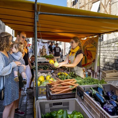 Une femme présentant des légumes à une femme portant sa petite fille, dans le marché de Blois.